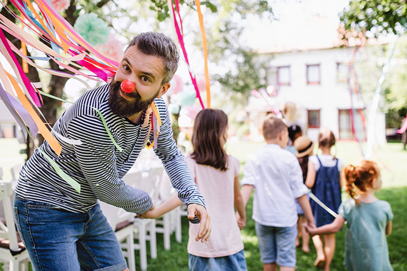 An adult wearing a clown nose is led by a group of children celebrating an event-planned birthday party.